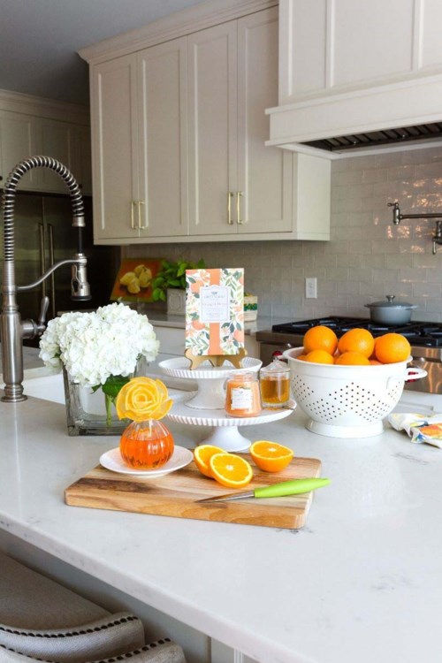 A kitchen counter with oranges, a glass of juice, flowers, and a cutting board. White cabinets and a stove are in the background.