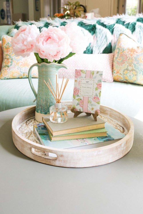 A tray on a table holds a vase with pink flowers, books, a reed diffuser, and a framed card. Cozy and decorative.