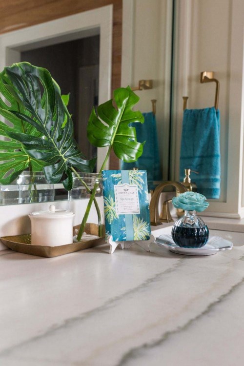 A bathroom counter with plants, candles, a decorative box, and a small glass bottle. Blue towels hang on the wall.