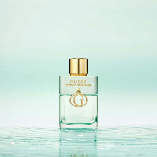 Een heldere parfumfles met gouden dop en GUESS-logo, drijvend op water met zachte golven en lichte achtergrond.