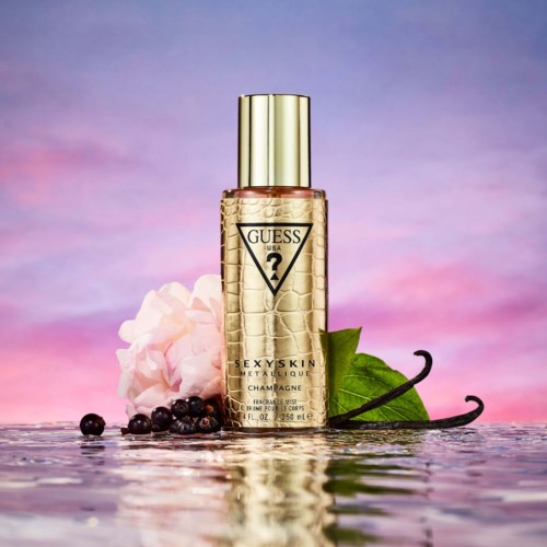 Een parfumfles met label 'Guess UGA 2 SEXY SKIN METALIC LOUNGE CHAMPAGNE' met roze bloemen, zwarte bessen, groen blad en spiegelreflexie.