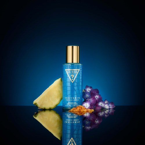 Een blauwe Guess-fles met gouden dop, ananas, druiven en bloembladeren voor, gereflecteerd op een donkere ondergrond.