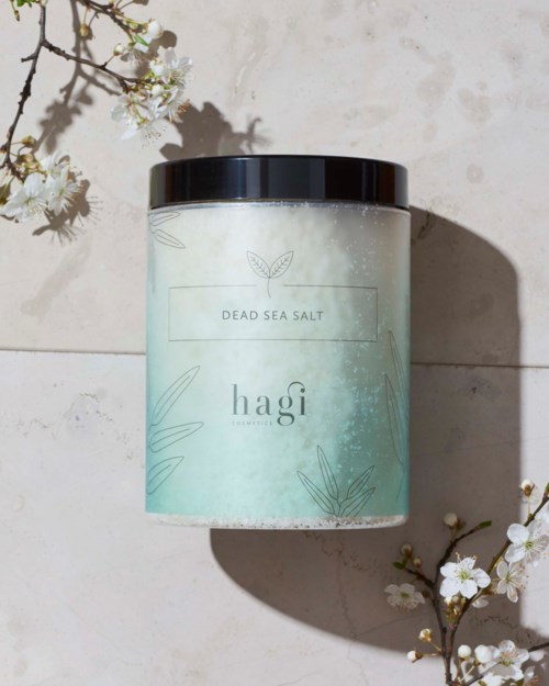 En behållare märkt 'Dead Sea Salt' på en ljus yta, omgiven av vita blommor med skuggor.