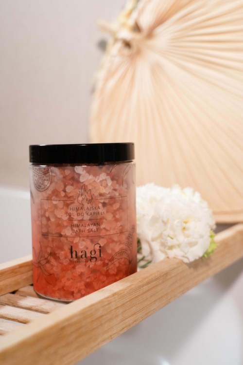 A jar with pink bath salts on a wooden shelf, with flowers and a decorative fan in the background.