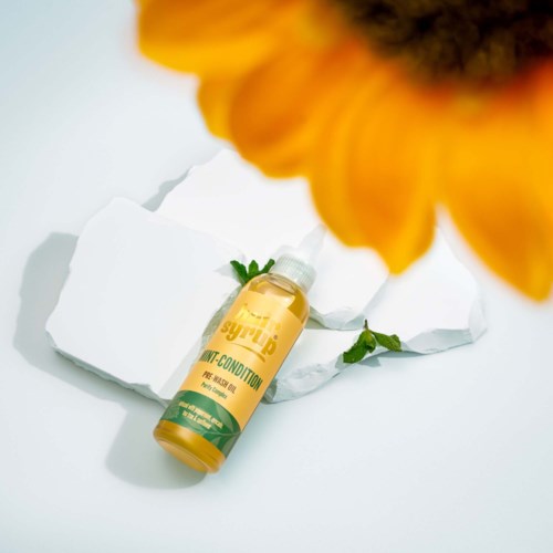 Een flesje oranje siroop haarconditioner ligt op gekreukt wit papier met een wazige gele zonnebloem op de voorgrond.