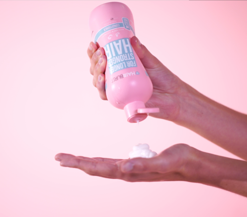 Hand die een roze fles vasthoudt, spuit schuim op handpalm tegen een roze achtergrond.