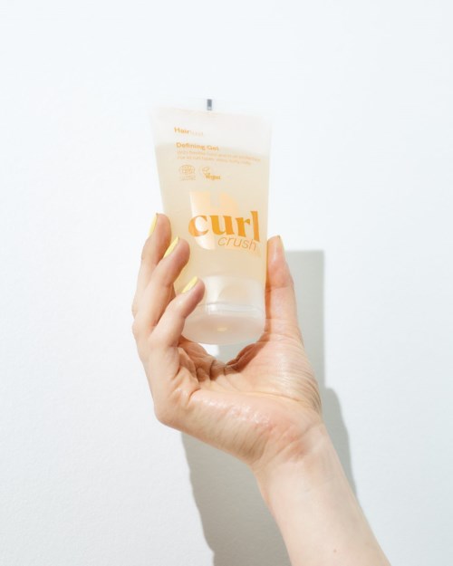 Hand holding a clear tube of curl defining cream with orange text and white cap.