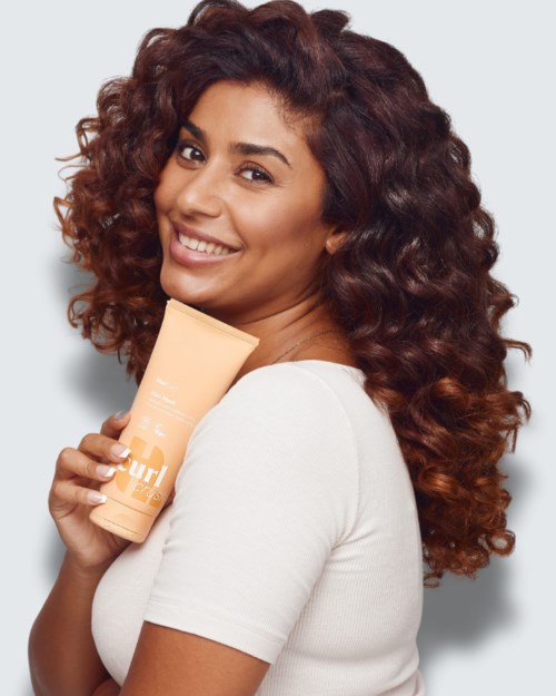 Smiling woman with curly hair holding a beige skincare tube, wearing a white top.