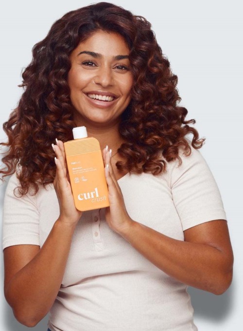 Glad kvinne som holder en flaske curl cream hårpleieprodukt, iført en hvit skjorte, med krøllete hår og grå bakgrunn.