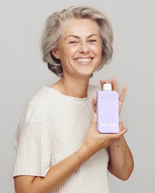 Smiling woman with gray hair holding a lavender bottle with blonde written on it.