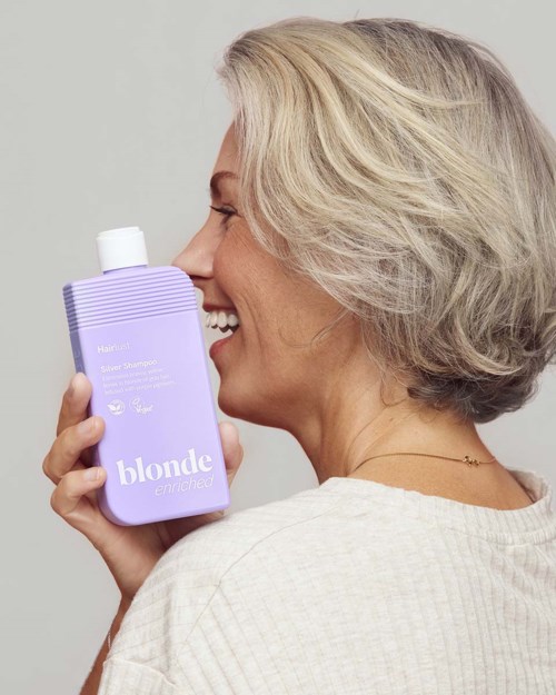 Smiling older woman with gray hair holding a purple shampoo bottle labeled blond. She is wearing a light-colored top.