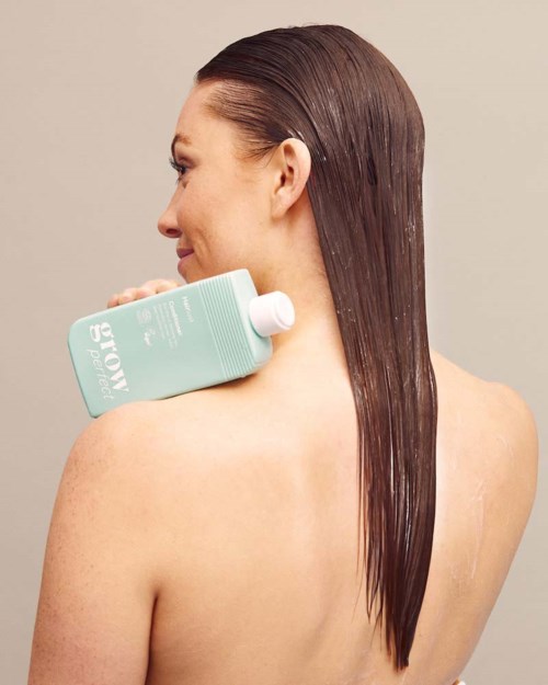Woman with long, straight brown hair holding a light blue shampoo bottle on her shoulder.