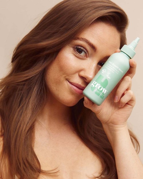 A woman with long brown hair holds a light green spray bottle close to her face, smiling gently.