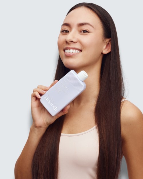 Happy woman with long dark hair holding a white skincare bottle, smiling towards the camera.