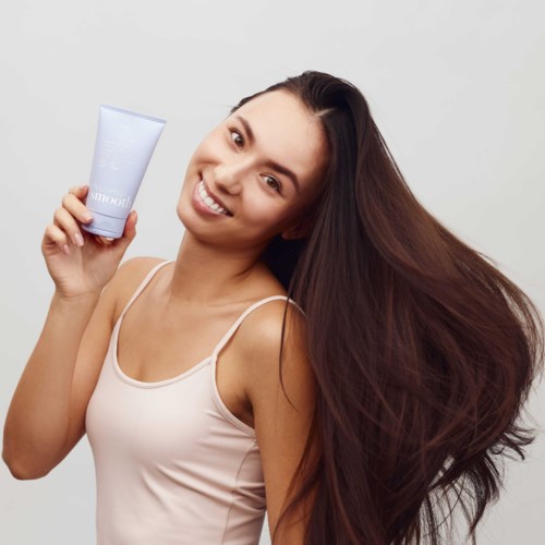 Smiling woman with long, brown hair holding a light blue skincare tube, wearing a beige tank top.