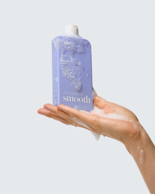 A hand holds a lavender shower gel bottle with foam on the surface and a mild lather. The label reads heavenly smooth.