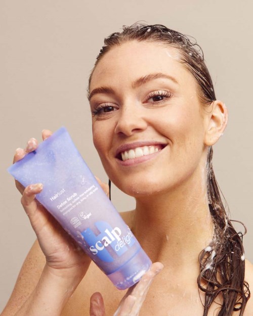 En kvinne smiler mens hun holder en blå tube med hårpleieprodukt kalt Scalp Delic.