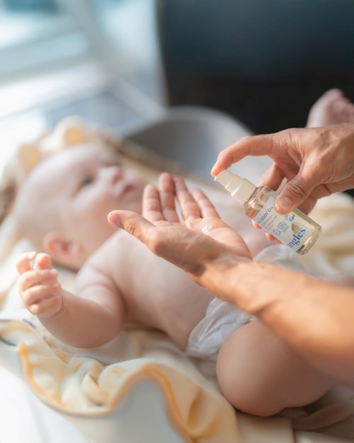 Person applying gentle skincare spray to a baby lying on a bed.