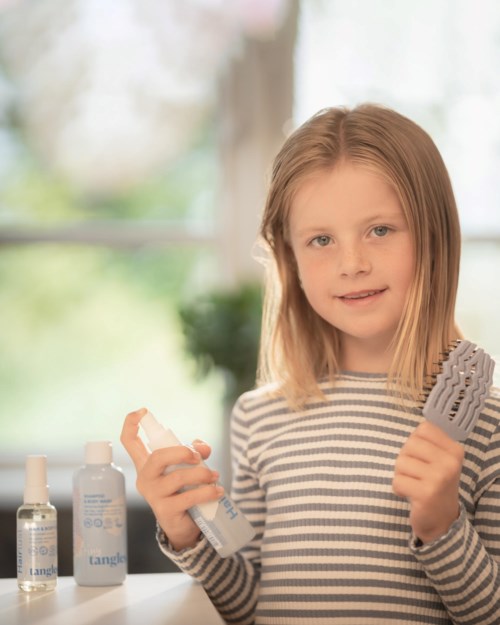 Young girl with blonde hair holding a hairbrush and a spray bottle, standing next to hair care products.