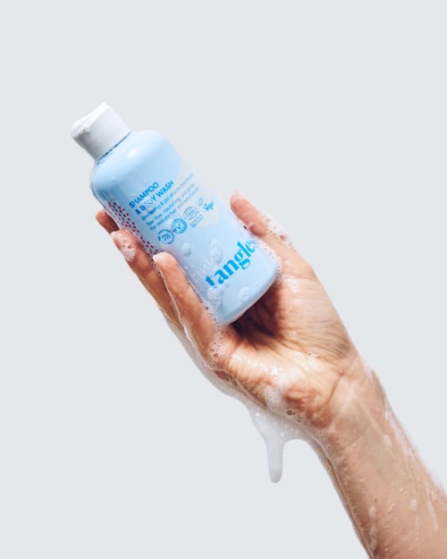 Hand holding a blue shampoo bottle with soap suds on hand, against a plain grey background.