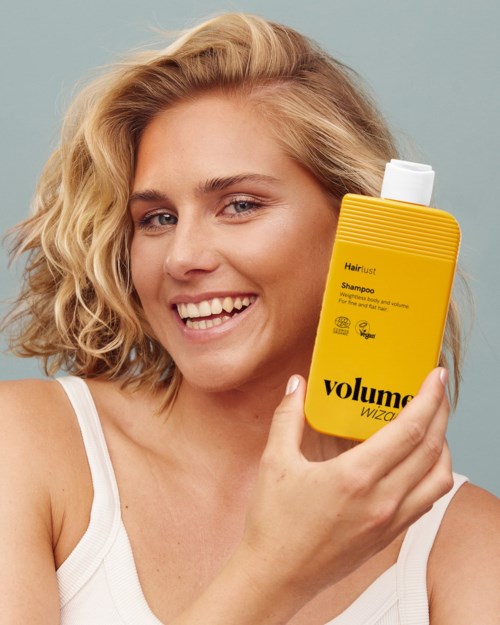 Smiling woman with short blonde hair holding a yellow shampoo bottle labeled Volume Wizard.