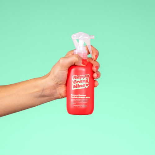 Eine Hand hält eine rote Sprühflasche mit der Aufschrift "Happy Crazy Minea" vor einem hellgrünen Hintergrund.