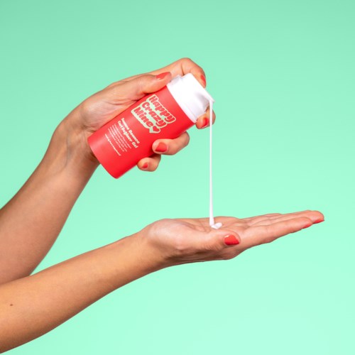 A person dispenses white lotion from a red bottle into their hand against a light green background.