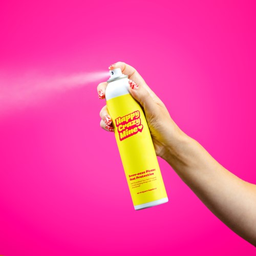 Een hand die een gele spuitbus vasthoudt met roze en rode tekst tegen een roze achtergrond.