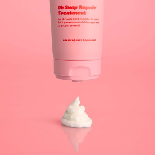 Een roze fles haarbehandeling met een klodder witte crème eronder tegen een roze achtergrond.