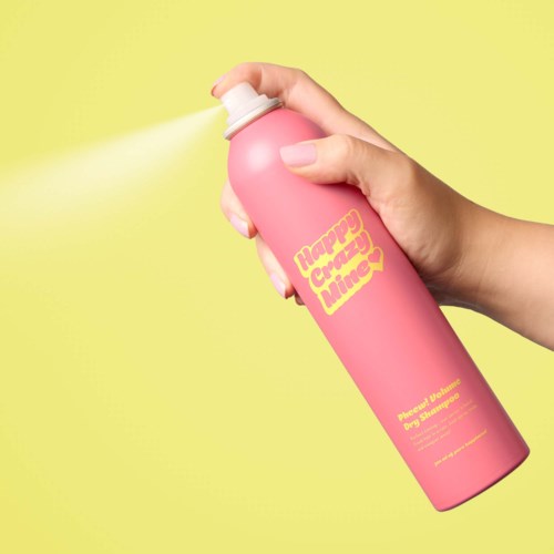 Een hand spuit roze haarlak met een gele label waarop staat "Happy Crazy Time" in gele tekst op een gele achtergrond.