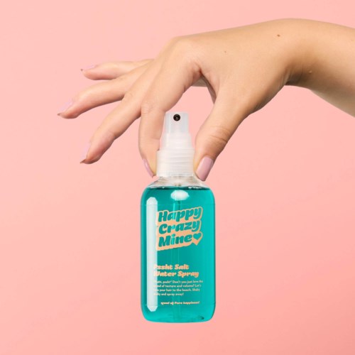 Een hand houdt een blauwe sprayfles met de tekst "Happy Crazy Mine" tegen een roze achtergrond.