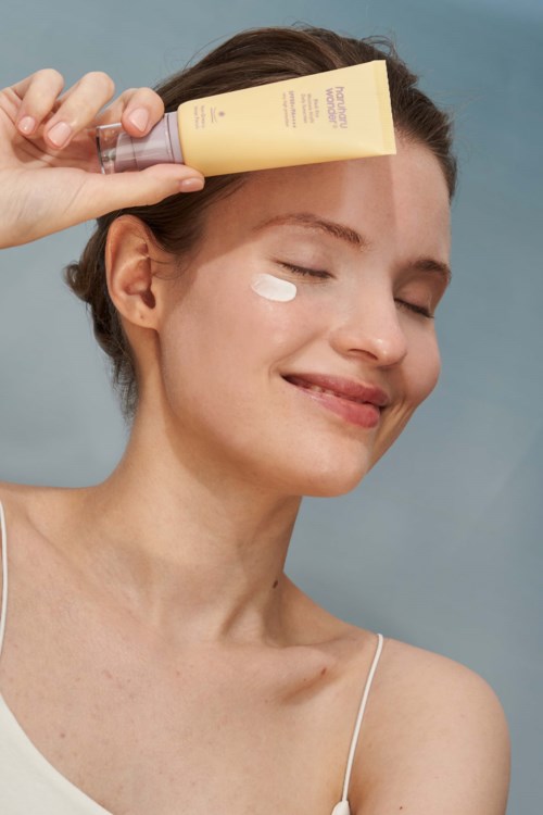 A woman smiles with eyes closed, applying lotion from a yellow tube on her face, against a blue background.