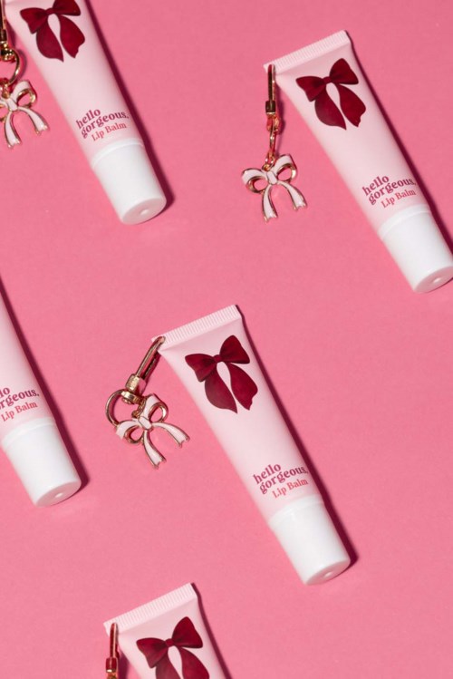 Små rosa leppepomade rør med røde sløyfer og små rosa sløyfeanheng, arrangert diagonalt på rosa bakgrunn.