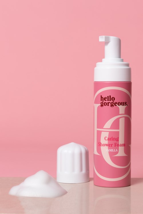 Een roze flesje met douche schuim, witte dop en een beetje schuim ernaast op een roze en grijze achtergrond.
