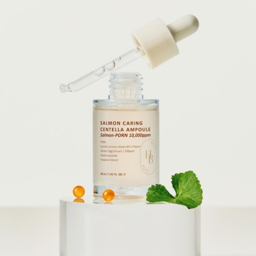 Eine Hautpflegeflasche mit Salmon Centella Ampulle, Tropfer, grünem Blatt und zwei orangefarbigen Pillen in schlichter Umgebung.