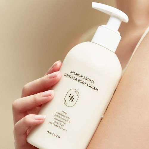 Eine Person hält eine weiße Flasche mit Lachs-Frucht-Body-Cream mit Pumpe, nahe der Schulter. Auf dem Etikett steht Text zu den Inhaltsstoffen.