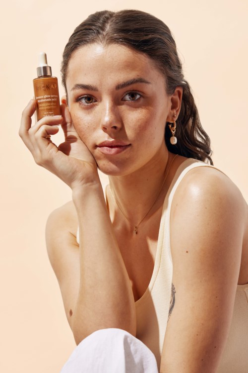 A woman holds a brown skincare bottle near her face, looking at the camera with a neutral expression.