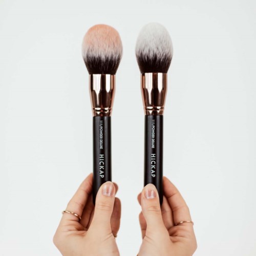 Two makeup brushes with black handles and pink and white bristles held by hands.