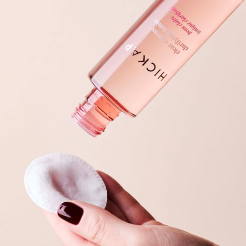 A hand holding a cotton pad with a bottle of HICKA skincare product pouring onto it.