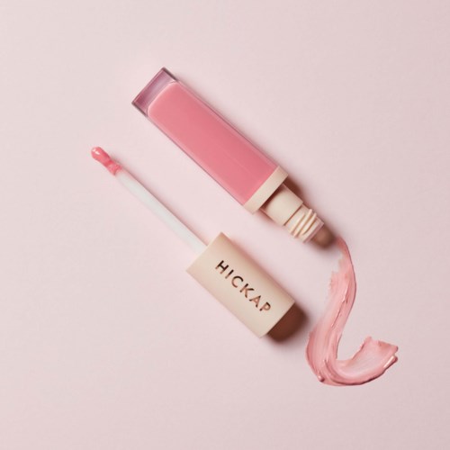 A pink lip gloss tube with the stick beside it, and gloss spread on a pink background.