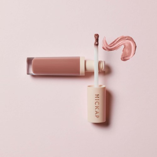 A lip gloss tube with its applicator outside, pinkish fluid spilled next to it on a pink background.