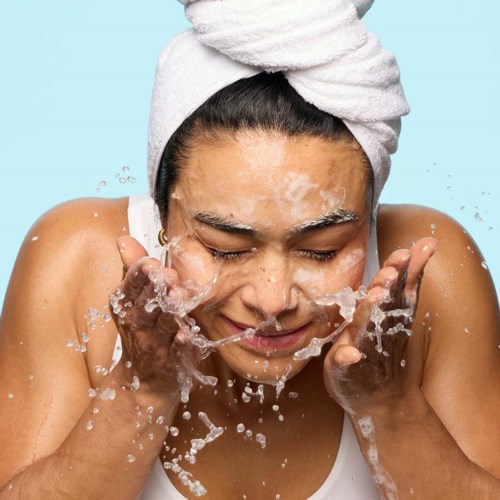 A woman with a towel on her head washes her face with water, smiling happily.