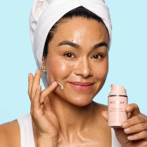 A woman with a towel on her head applies skincare cream to her face, smiling. She holds a pink bottle.