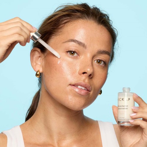 A woman applies serum to her face using a dropper, holding a small bottle of skincare product.