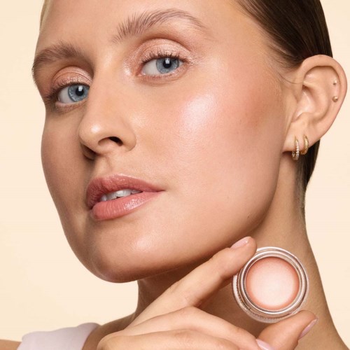 A woman with clear skin and blue eyes holds a small cosmetic jar near her face.