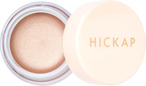 A small round container with a beige, shimmery makeup product and a white lid with gold letters saying "HICKAP."