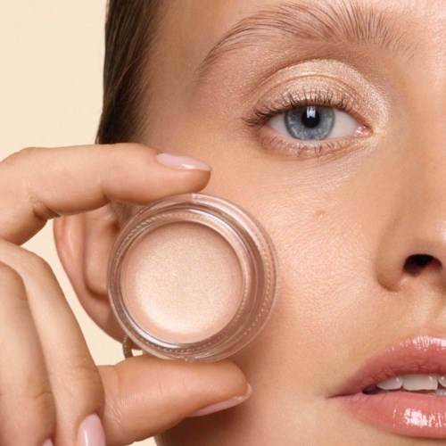 Close-up of a woman applying light-colored eyeshadow with her finger. Her face is partially visible with clear skin and light lipstick.
