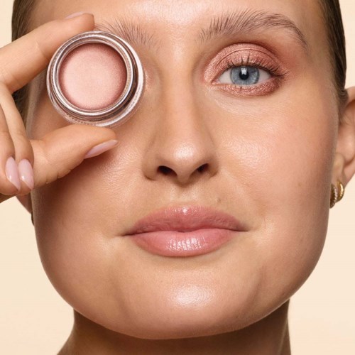 A woman holds a small jar of pink blush near her eye, smiling slightly with clear, glowing skin and blue eyes.