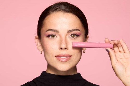 A woman with pink makeup holds a pink object near her nose against a pink background.