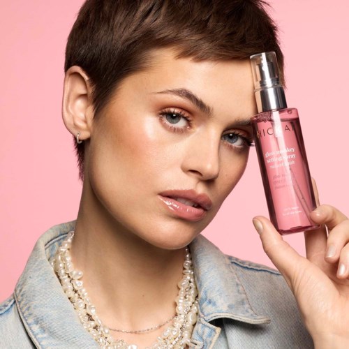 A woman with short hair holding a pink skincare bottle near her face, against a pink background.